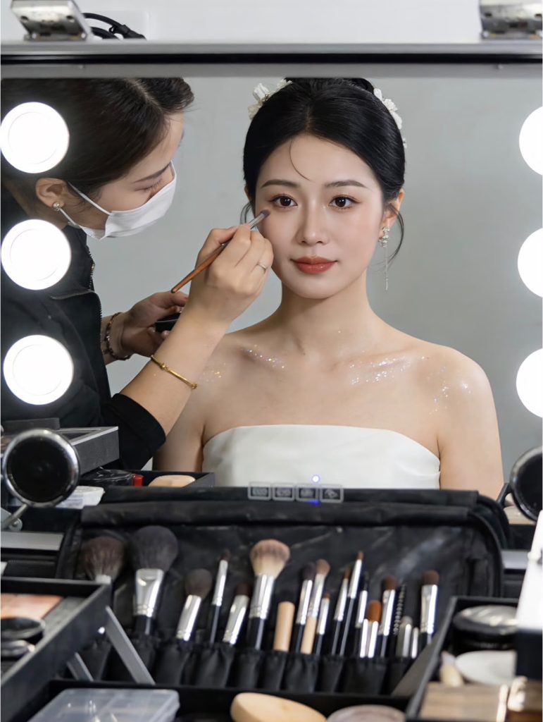 A makeup artist brushing eyeshadow onto a client in bridal hair and makeup at a lit vanity mirror