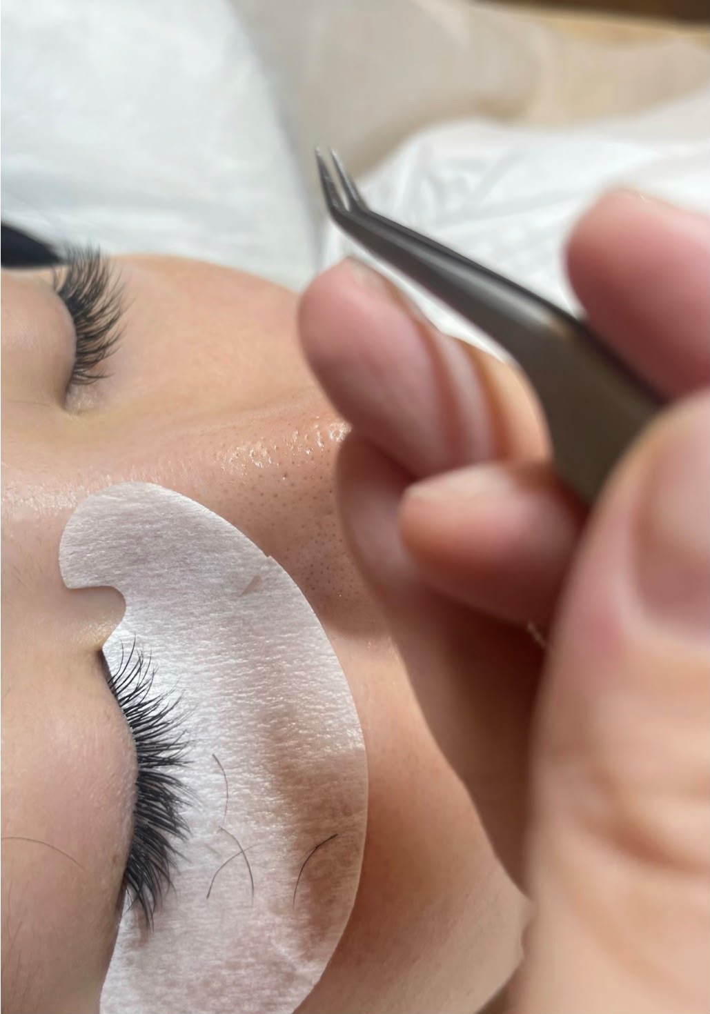 A lash artist using fine tweezers to apply extensions, with an under-eye pad protecting the lower lashes